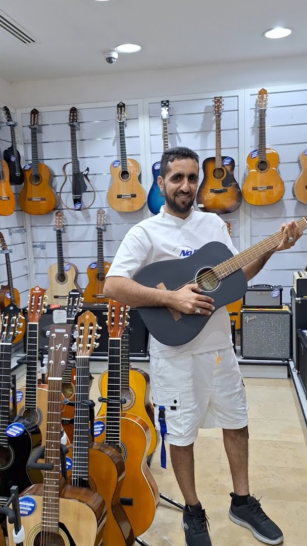 A man holds a guitar in a music shop filled with various guitars and amplifiers.