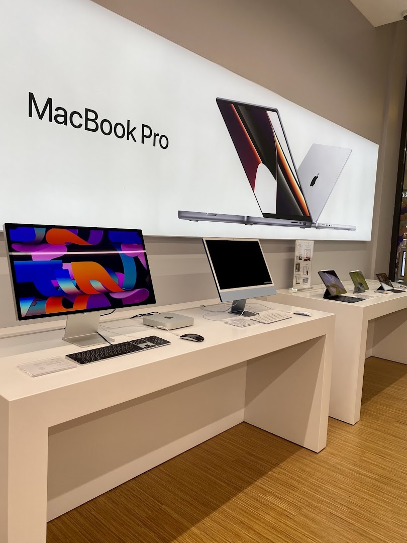 Display of Apple MacBook Pro and iMac computers on white tables in a retail store.
