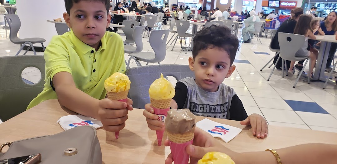 Two young boys enjoy ice cream cones at Baskin Robbins in a busy mall food court.