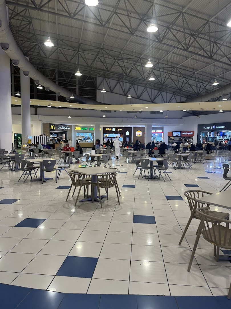 A food court with multiple restaurants and seating areas under a large, industrial-style ceiling.