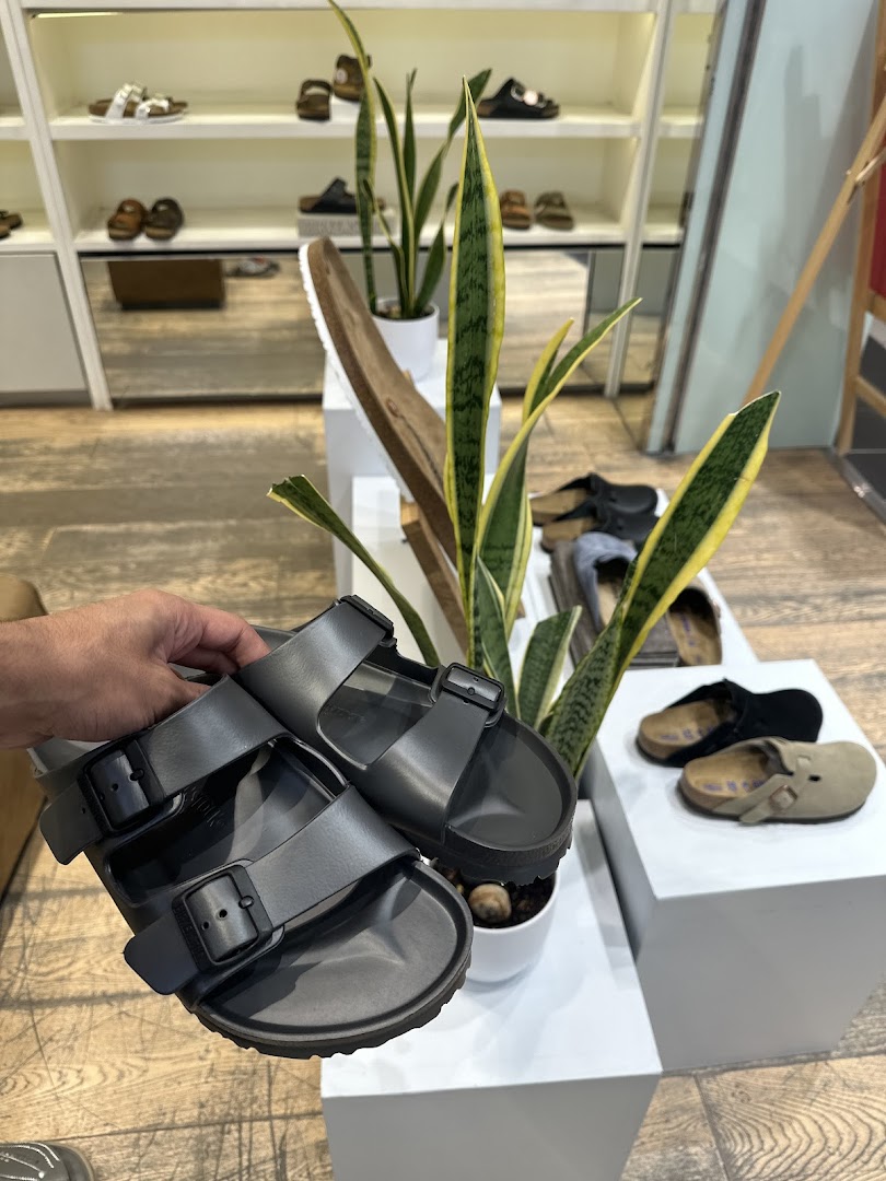 A hand holds a pair of dark grey Birkenstock sandals in front of a display of shoes and plants.