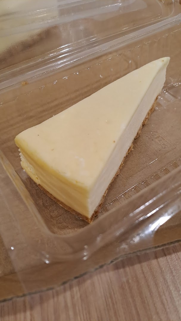 A slice of creamy cheesecake in a clear plastic container on a wooden surface.