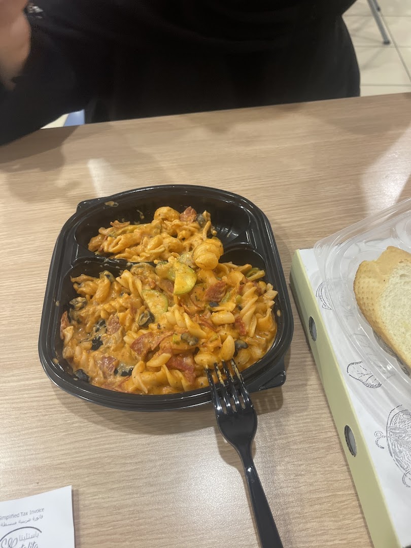 A close-up of a pasta dish with vegetables and a slice of bread on a wooden table.