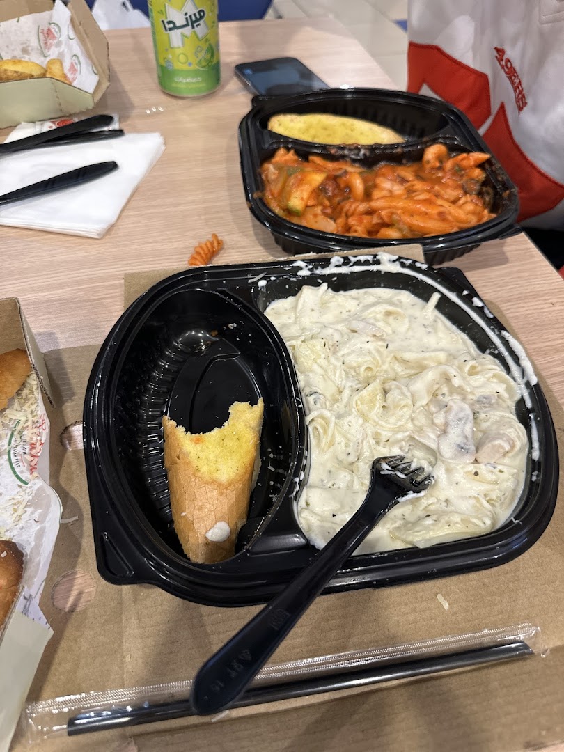 Two takeaway pasta dishes with garlic bread and a drink on a table.