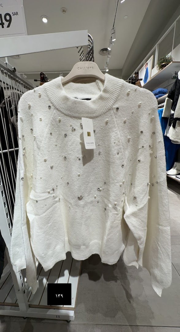 A white Calliope sweater adorned with pearls and rhinestones, displayed on a hanger.