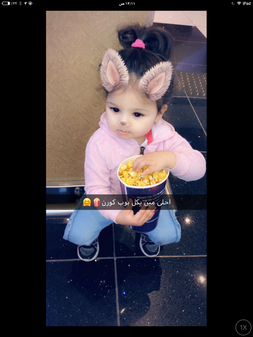 A young child with cat ear filters holds a large bucket of popcorn.