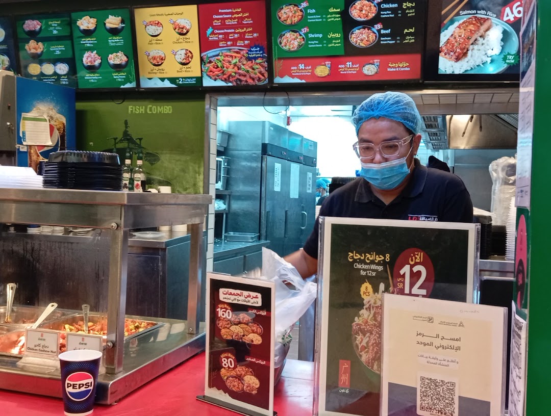 A restaurant counter with a menu board, food displays, and a staff member wearing a mask and hairnet.