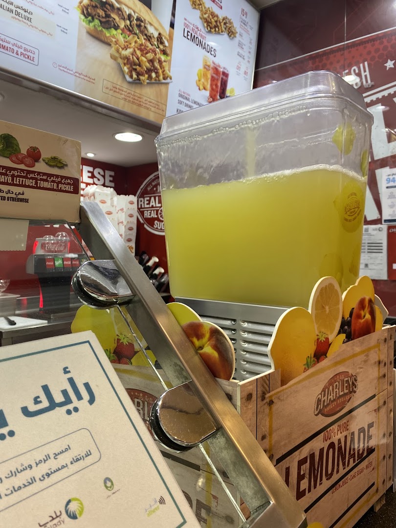 A large dispenser of lemonade is shown with a display of fruit and a Charleys Cheesesteaks sign.