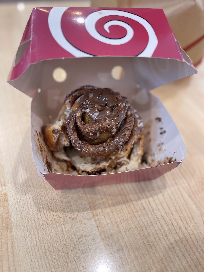 A close-up of a Cinnabon cinnamon roll with chocolate frosting in a branded box.