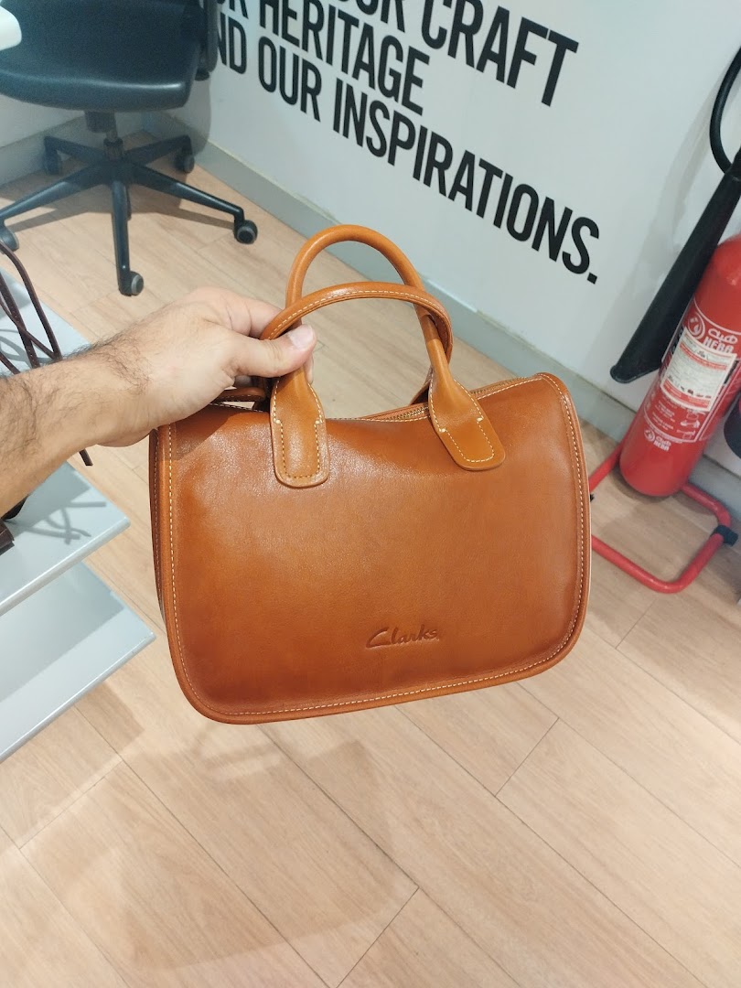 A hand holds a brown leather handbag with the Clarks logo embossed on it.