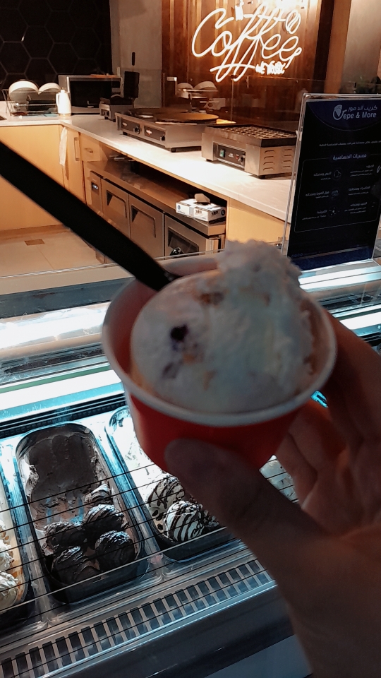 A hand holds a cup of ice cream in front of a display case with various flavors.
