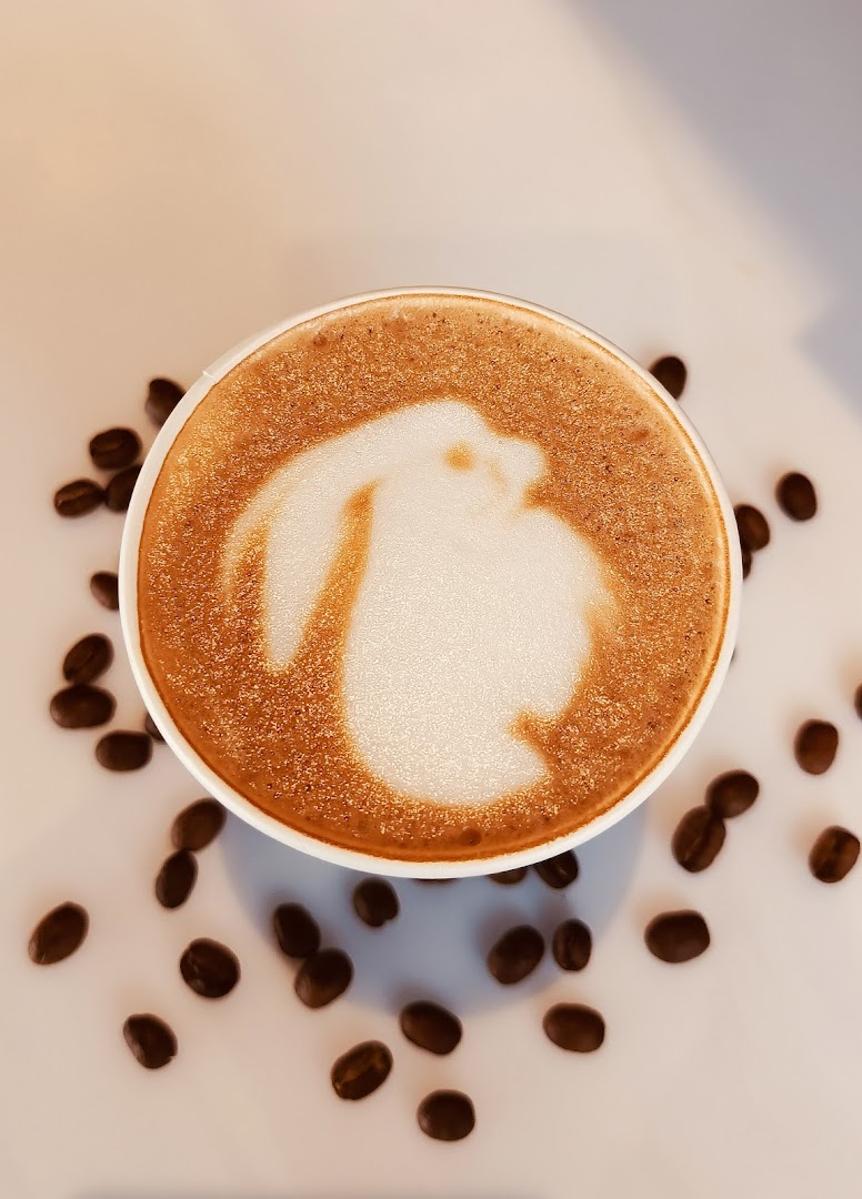 A cup of coffee with latte art resembling a rabbit, surrounded by coffee beans.