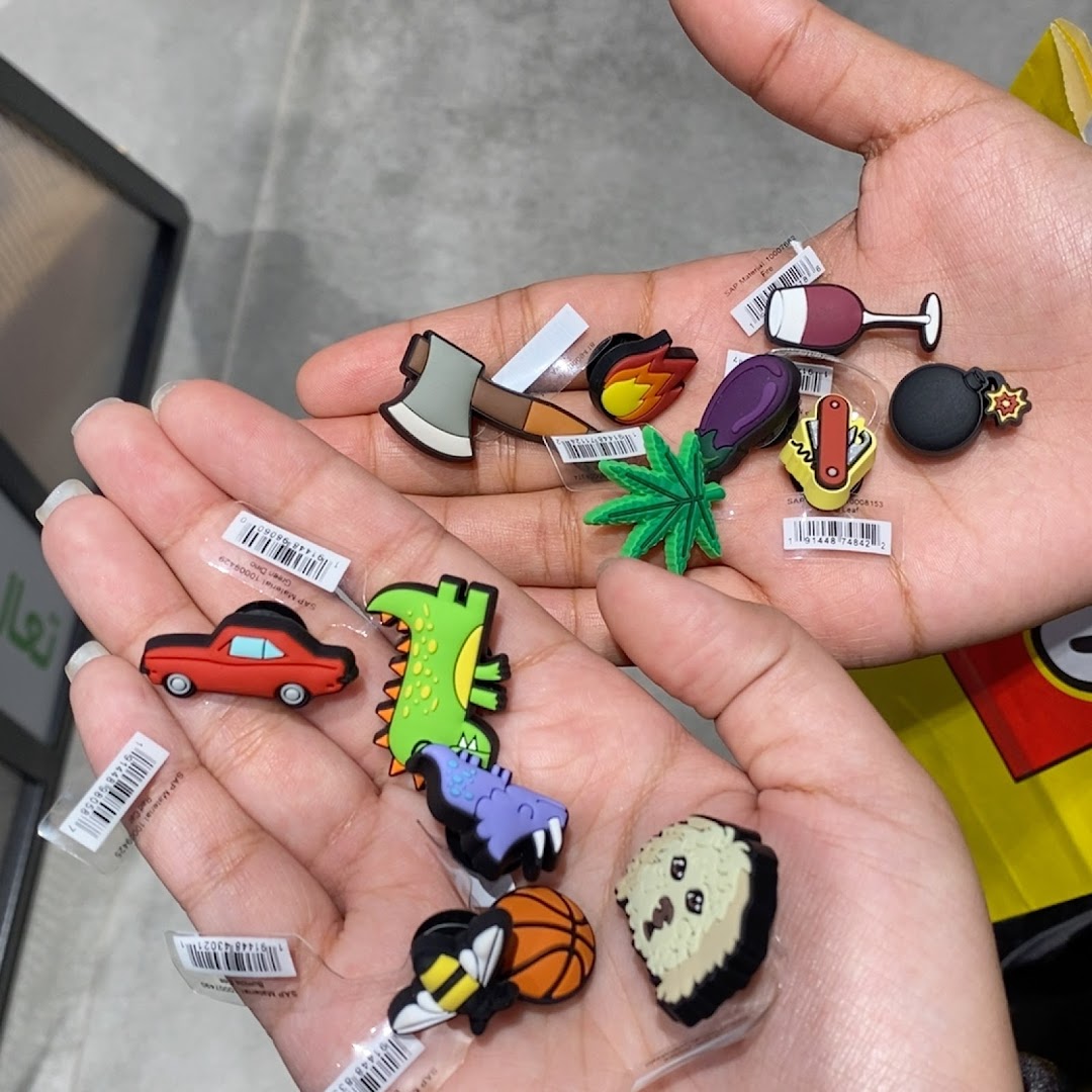 Hands holding various colorful Crocs Jibbitz charms including a dinosaur, car, axe, and bee.