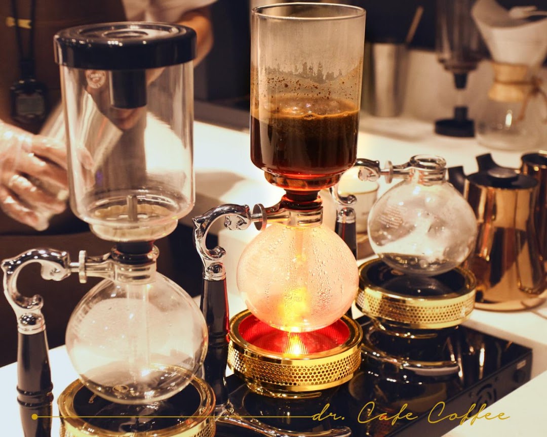 Close-up of a siphon coffee maker with brewing coffee and a barista in the background.