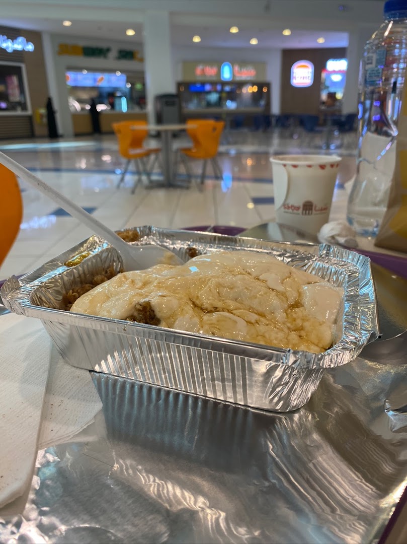 A foil container of food with a creamy topping and a spoon in a mall food court.