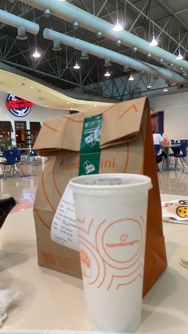 A Hamburgini paper bag and branded cup sit on a table in a food court.