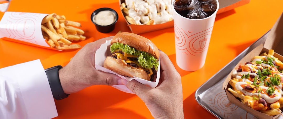 A person holds a double-patty burger with lettuce and cheese, next to fries and a drink.