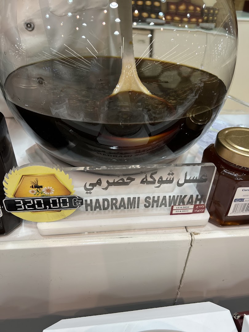 A large glass container filled with dark honey, with a sign indicating 'Hadrami Sawkah' and a price.