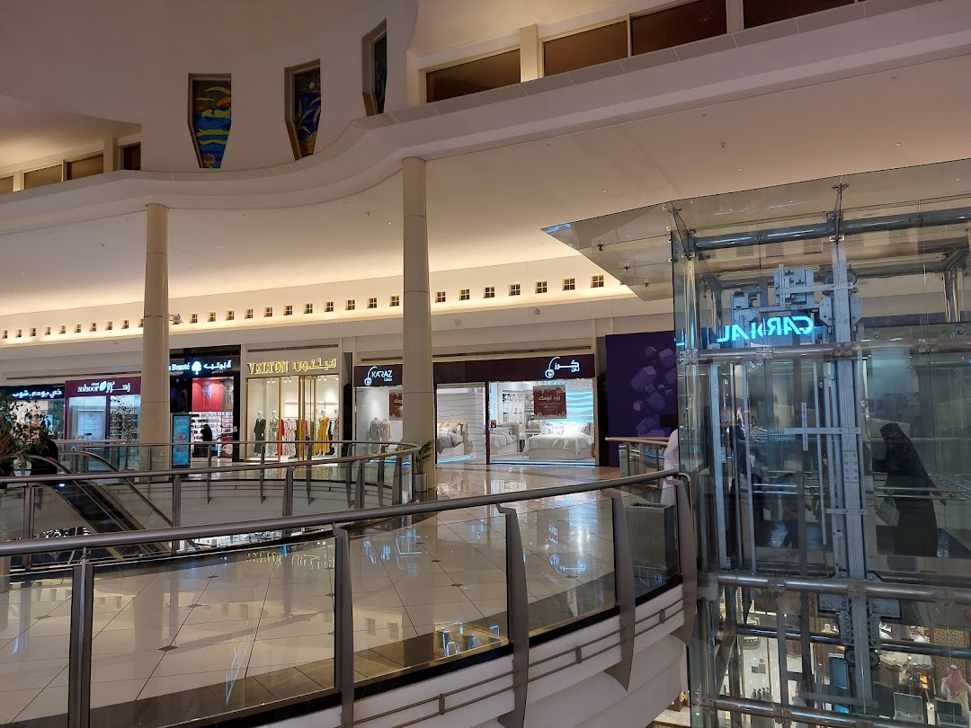 A view of a shopping mall interior with various shops, including Karaz Linen, and a glass elevator.