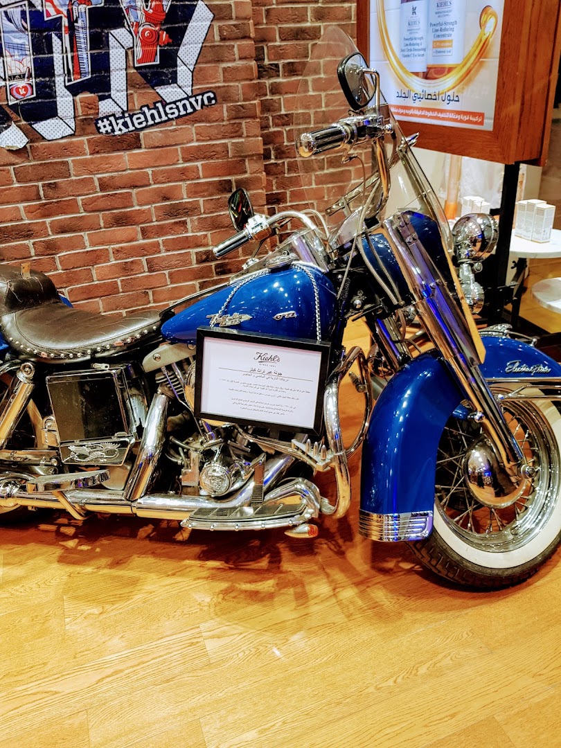 A blue motorcycle is displayed in front of a brick wall with Kiehl's branding.