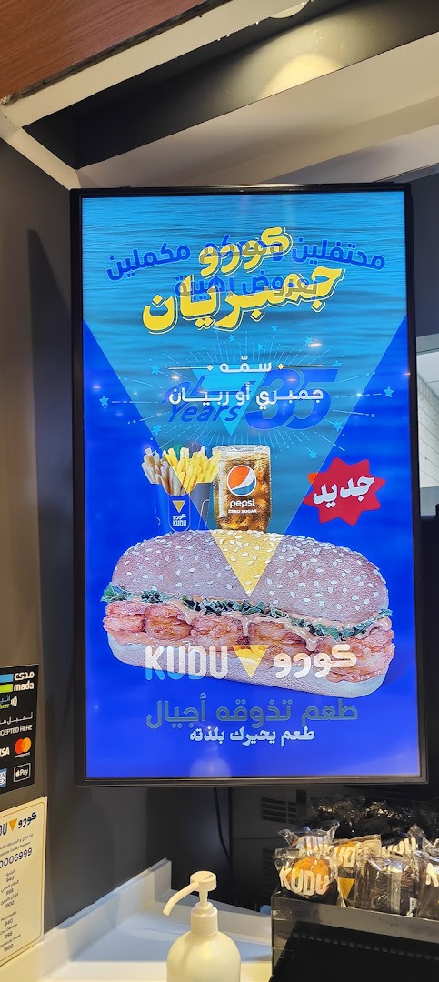 A digital menu board displays a large shrimp burger, fries, and a Pepsi, with Arabic text promoting the restaurant.