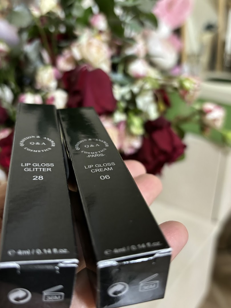 Two boxes of Q&A Cosmetics lip gloss, one glitter and one cream, held in front of flowers.
