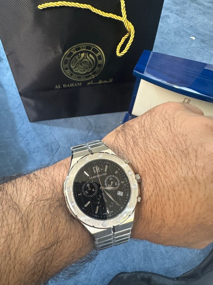A person wearing a silver watch with a black dial, next to an ALDAHAM shopping bag and a blue watch box.