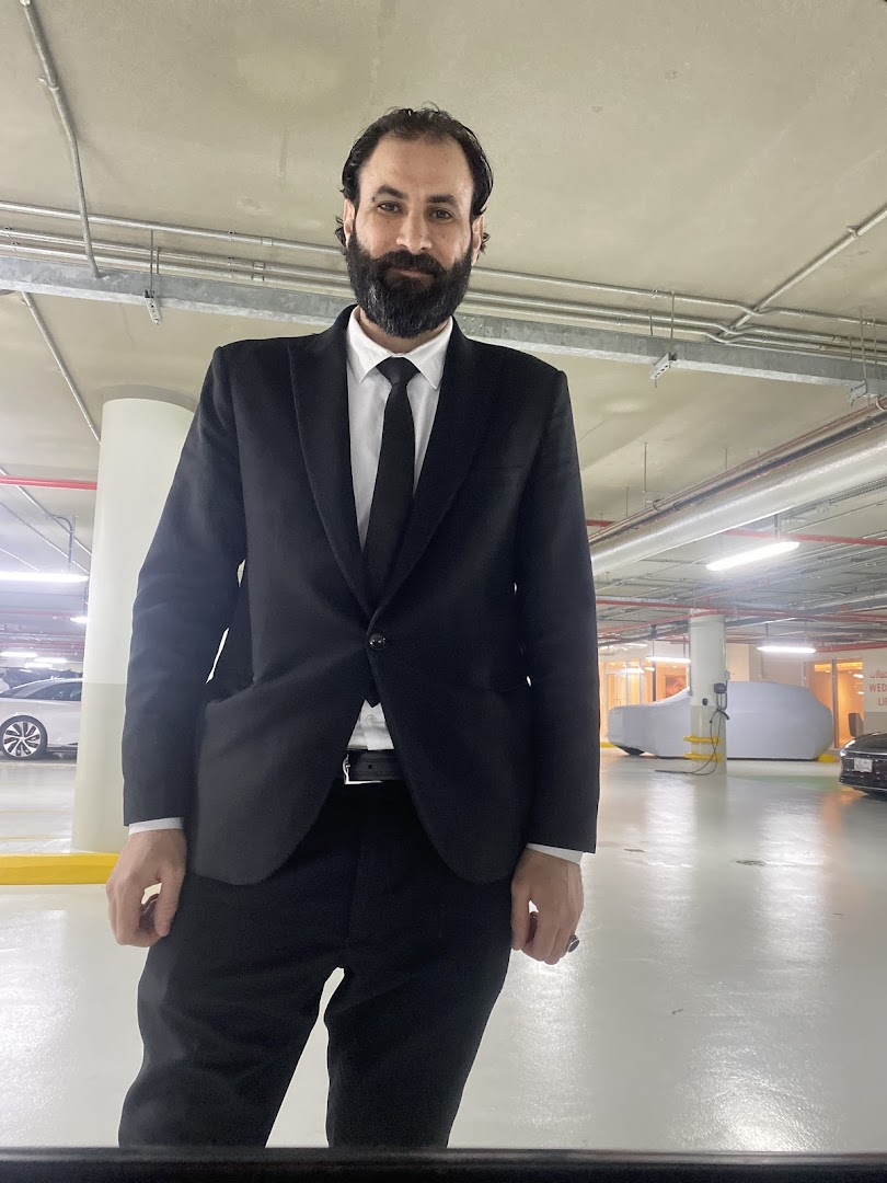 A man in a black suit and tie stands in a well-lit parking garage.