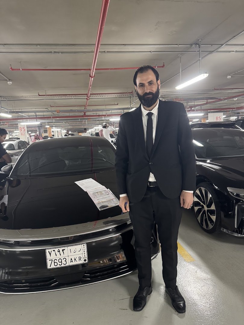 A man in a suit stands in front of a luxury car in a parking garage.