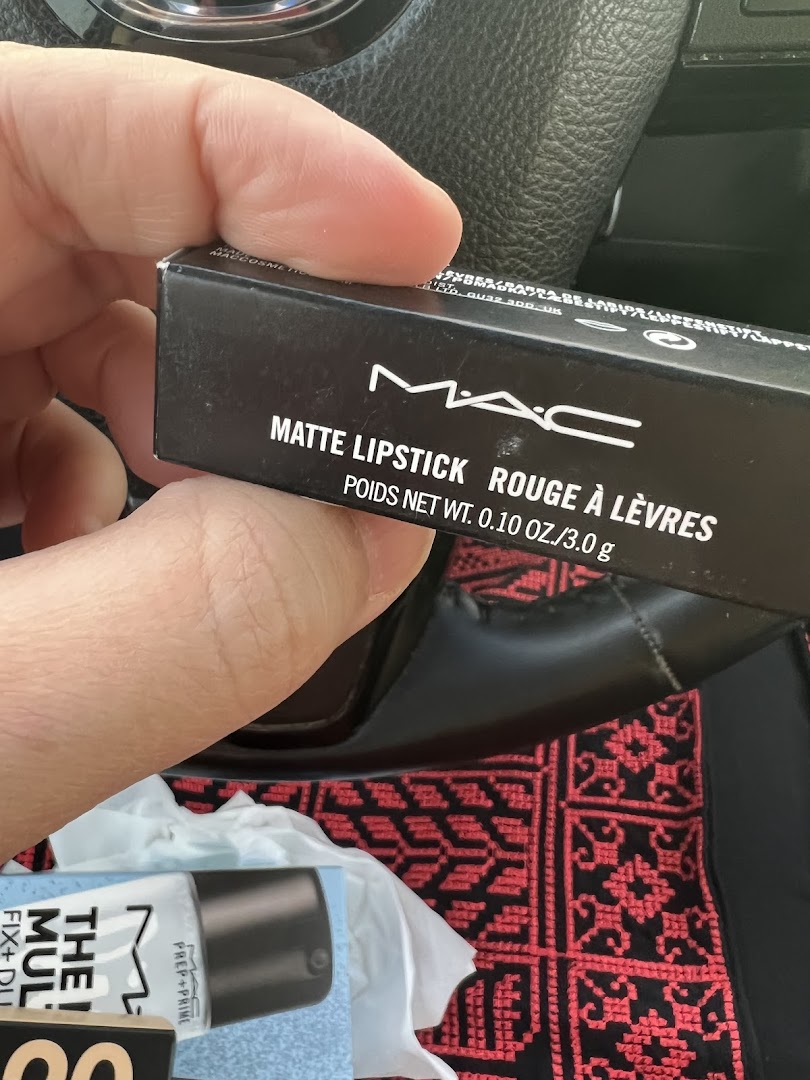 A hand holds a MAC Matte Lipstick box with other MAC products in the background.