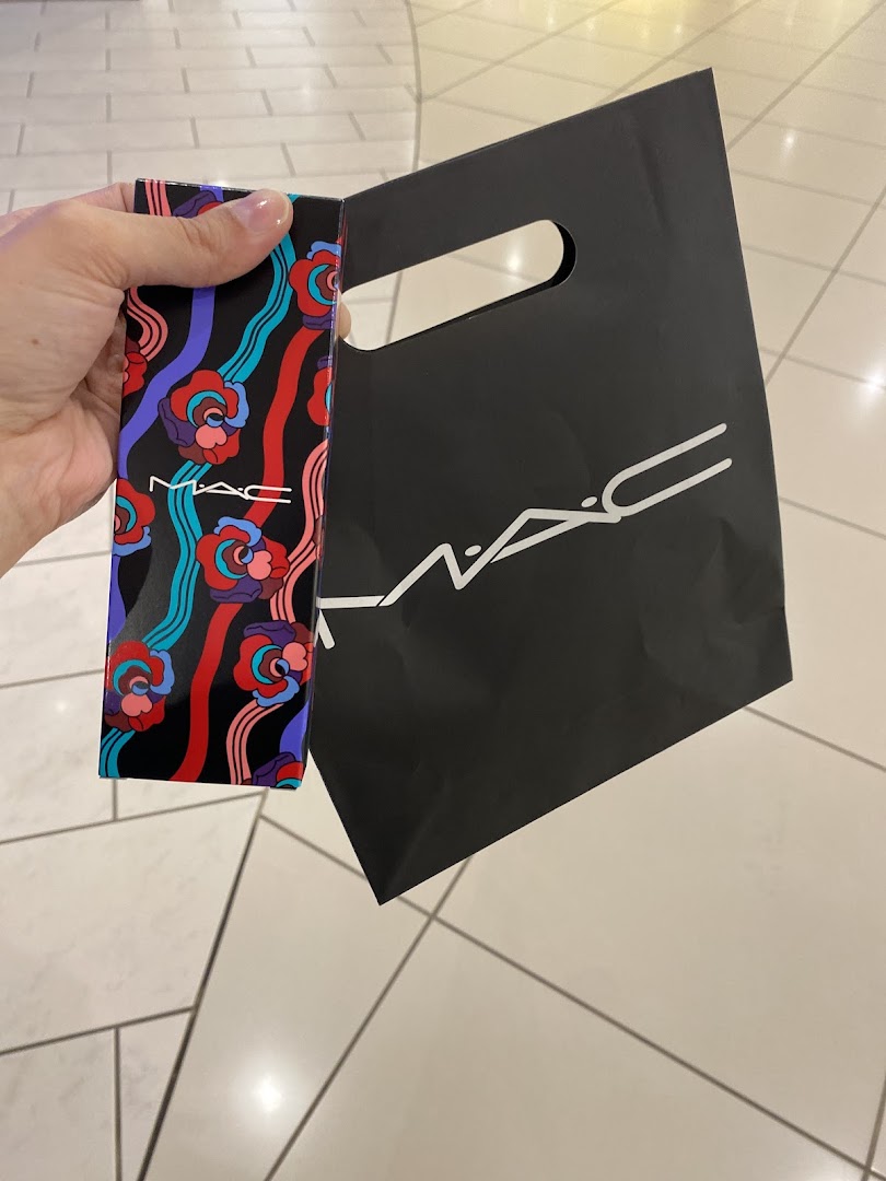 A hand holds a colorful M.A.C. product box next to a black M.A.C. shopping bag.