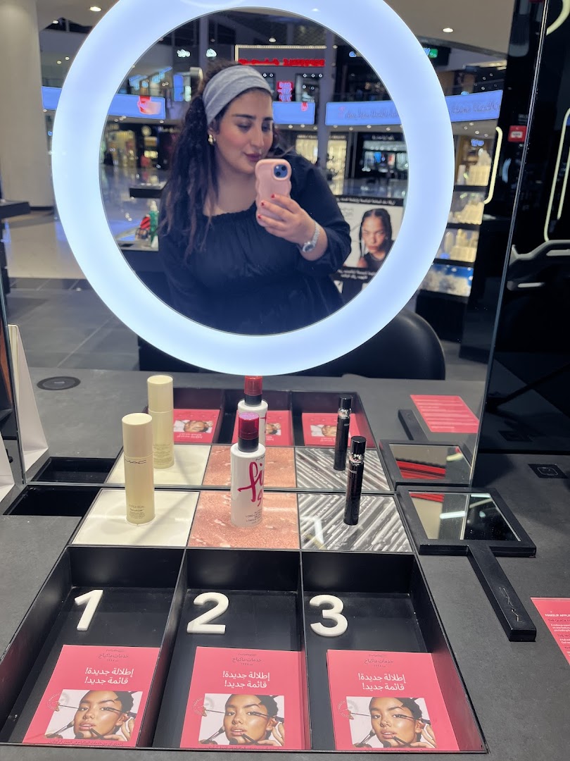 A woman takes a selfie in a M.A.C. store, reflected in a ring light, with makeup products displayed.