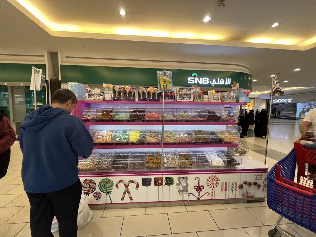 A man browses a colorful candy display in a mall, with other shoppers and store signs in the background.