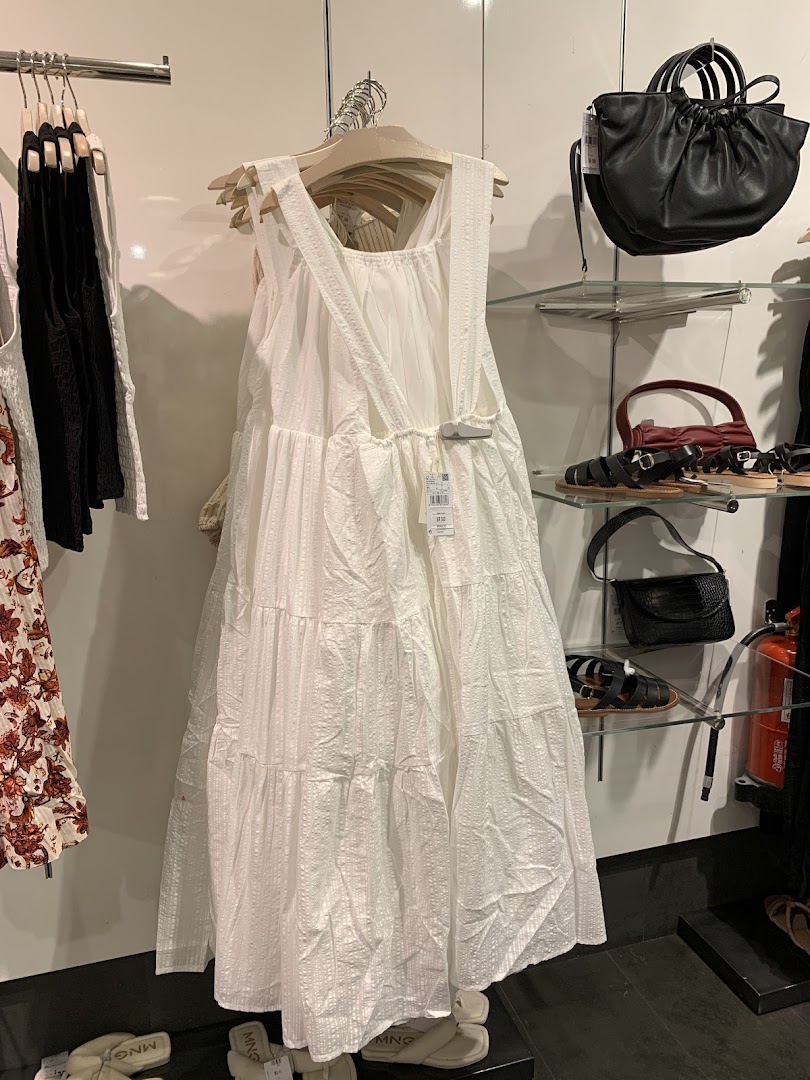 A white tiered dress is displayed on a hanger, with handbags and sandals on shelves.
