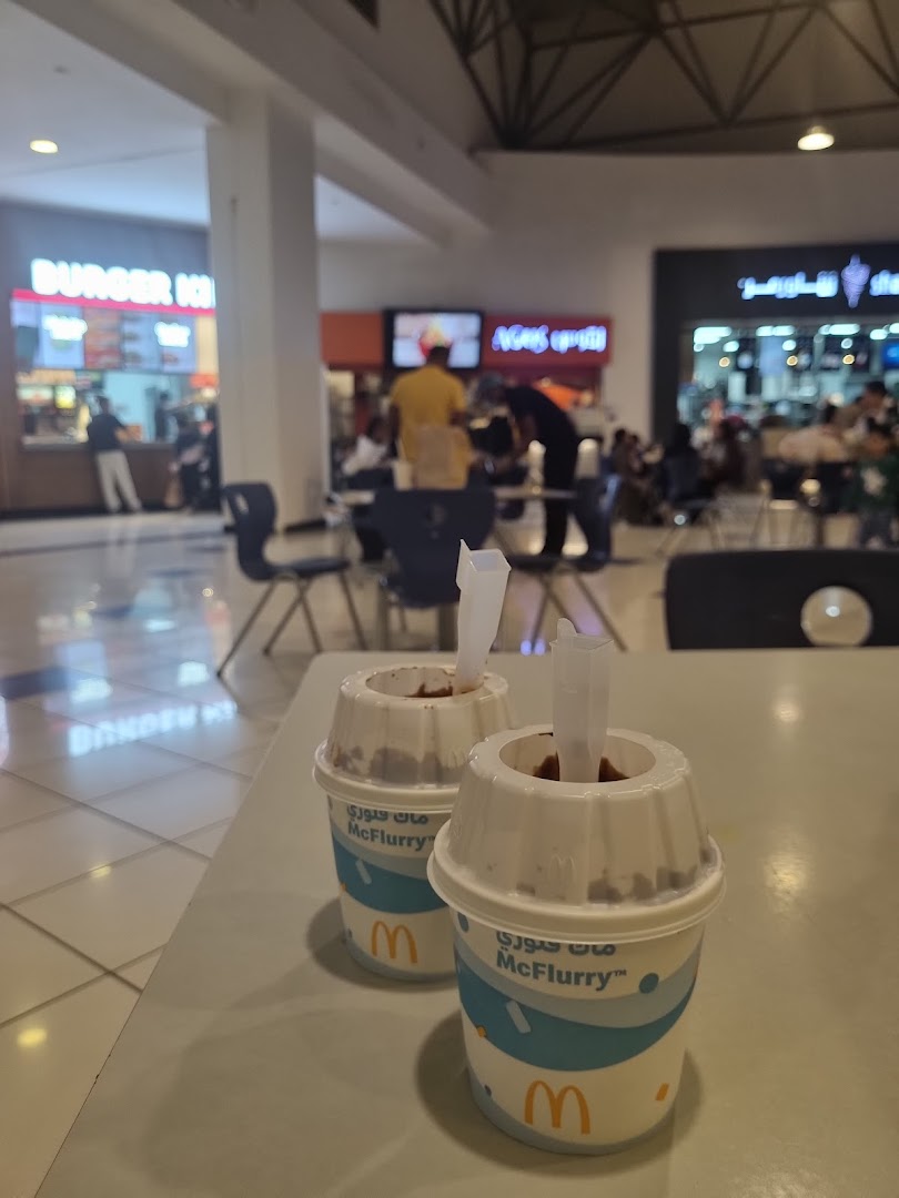 Two McFlurry desserts are placed on a table in a food court with other restaurants in the background.
