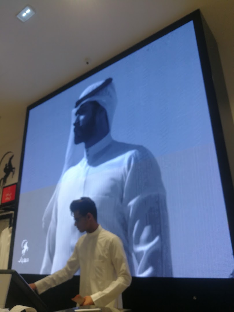 A man in traditional attire stands in front of a large screen displaying a similar figure.