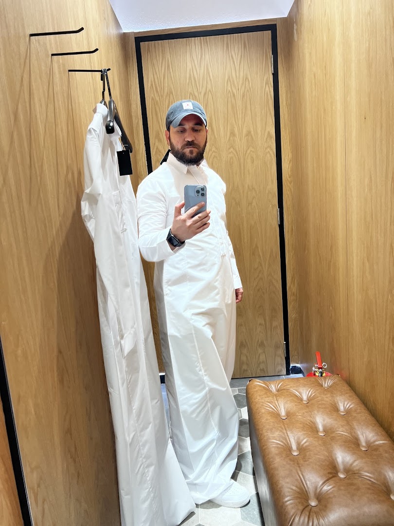 Man in a white thobe and cap taking a selfie in a fitting room.