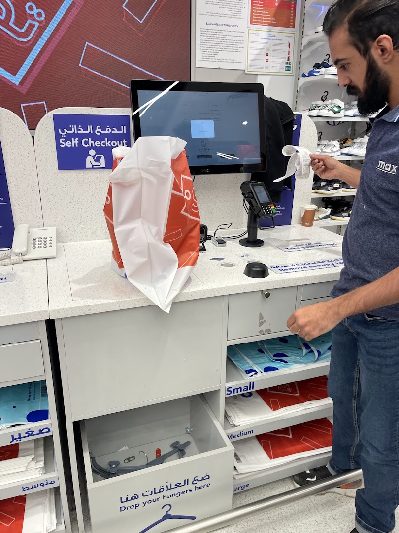 A man uses a self-checkout counter at a Max store, with shopping bags and a receipt.
