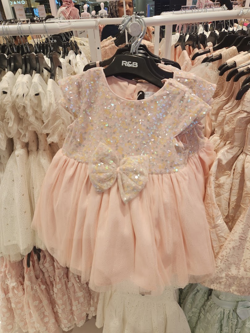 A close-up of a pink sequined baby dress with a large bow, displayed on a rack with other dresses.