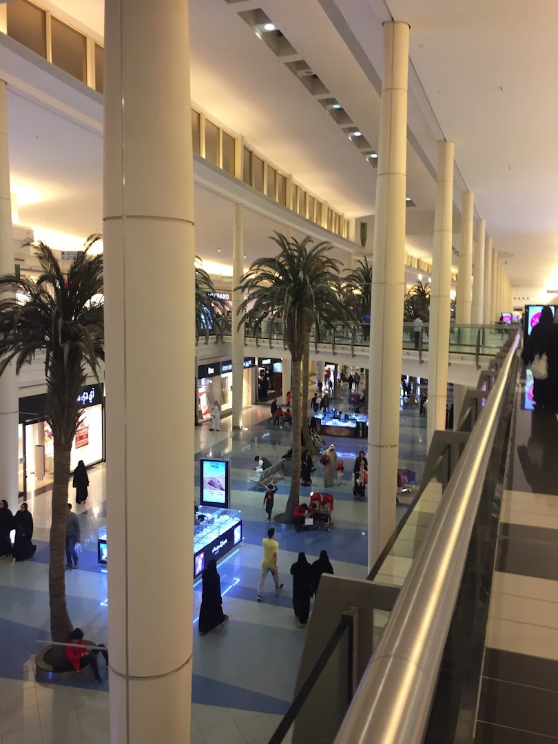 Interior view of a shopping mall with palm trees, shops, and people walking around.