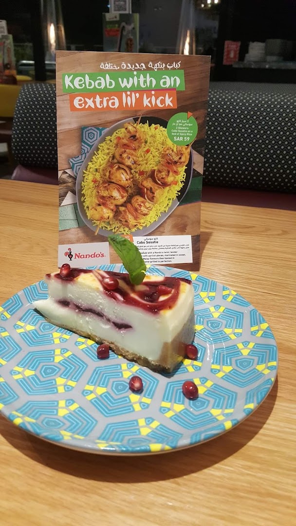 A slice of cheesecake with berry swirl and pomegranate seeds on a decorative plate, with a Nando's menu in the background.
