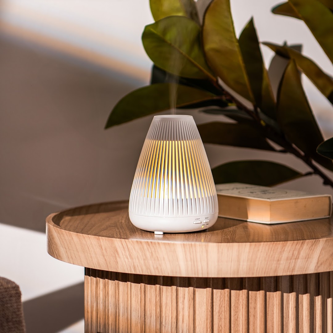 A white aroma diffuser with yellow light sits on a wooden table next to a book.