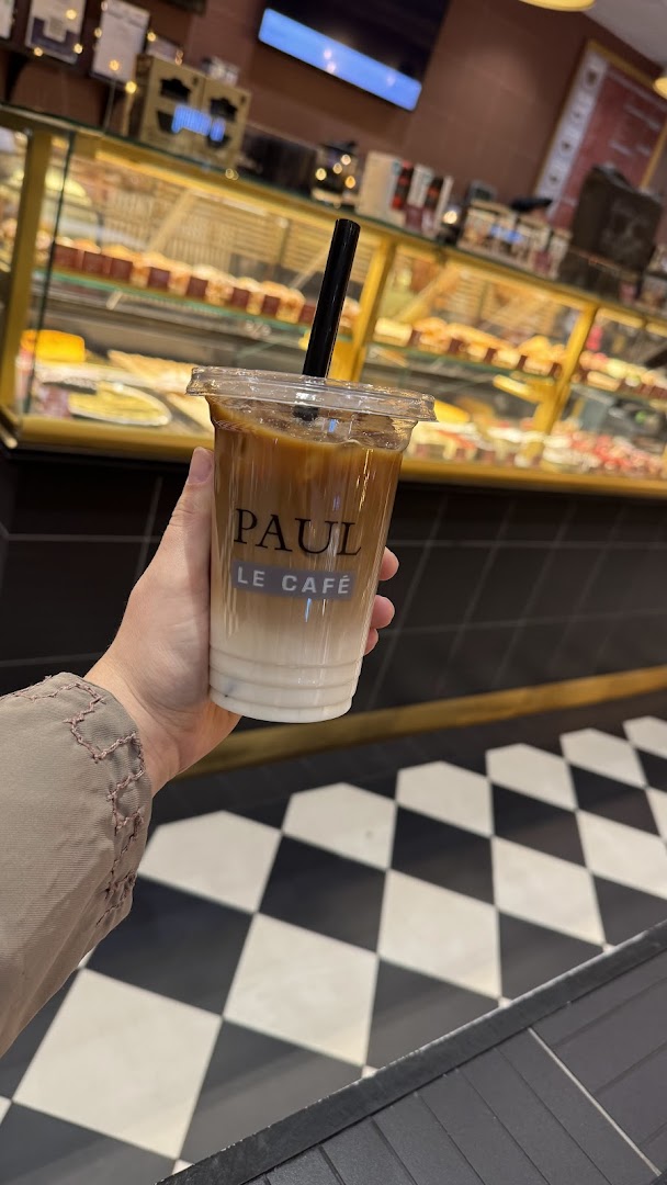 A hand holds a layered iced coffee from Paul Le Cafe in front of a display of pastries.