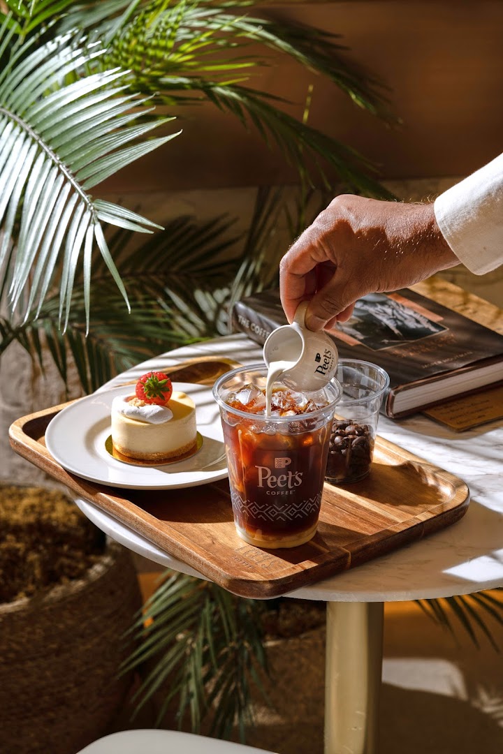 A hand pours cream into an iced coffee next to a small cake and coffee beans.