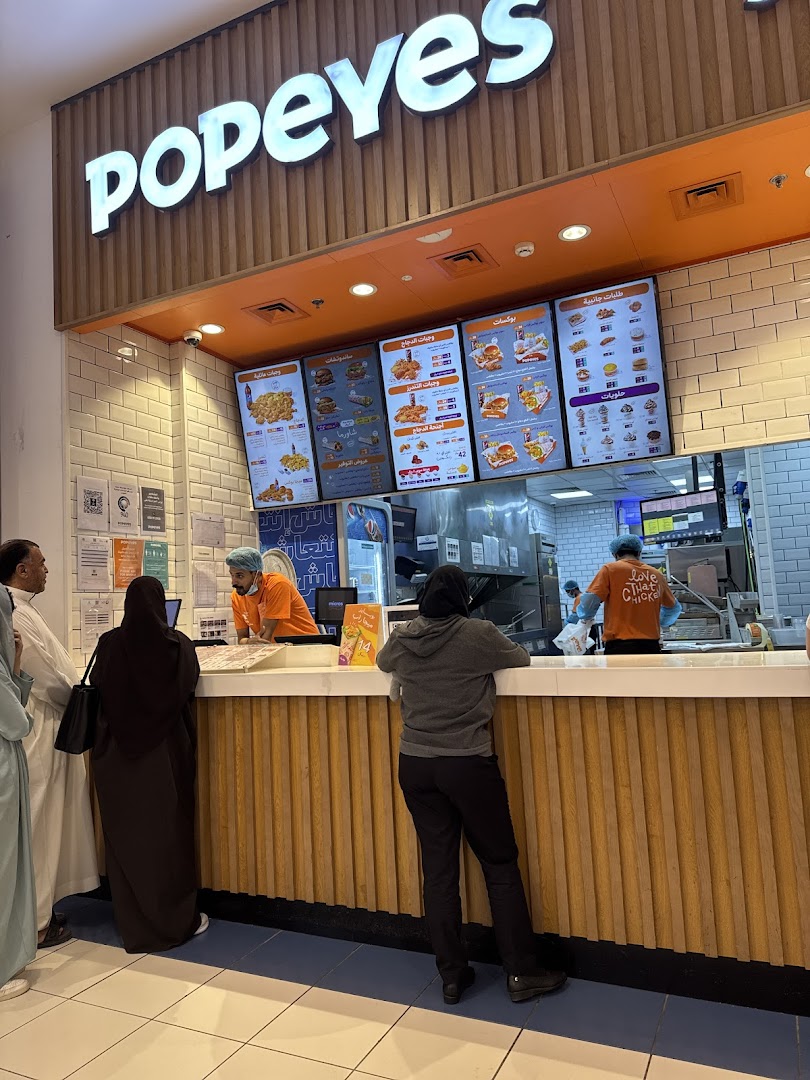 Customers ordering at the Popeyes counter with digital menus displaying food items.