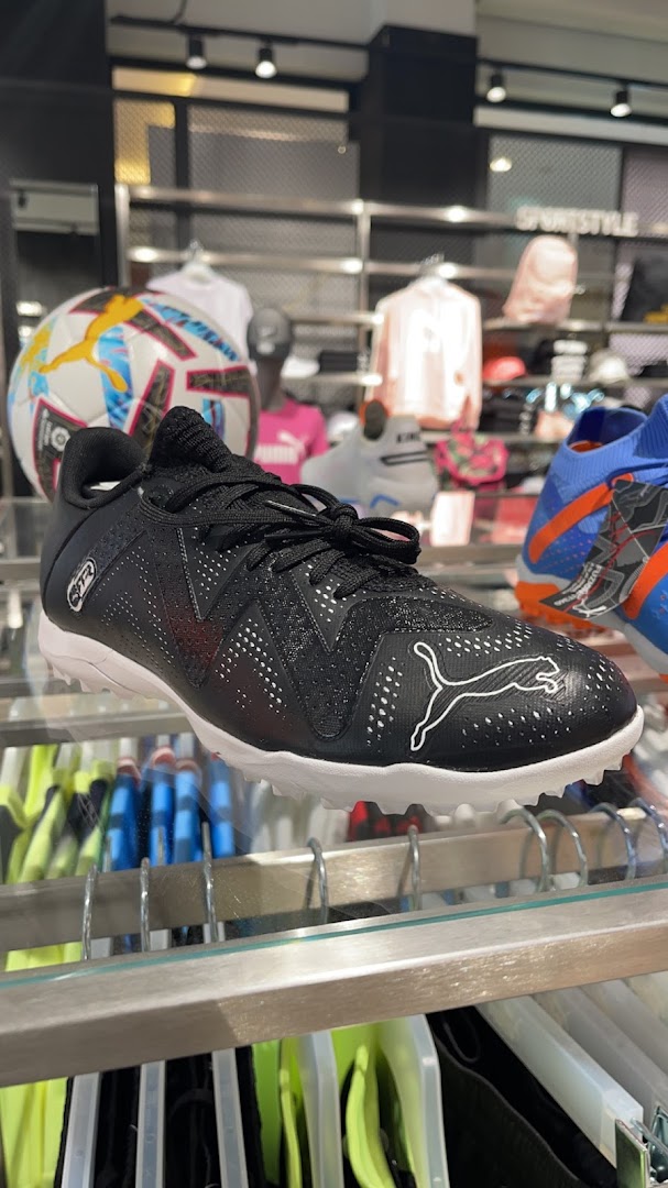 A black Puma soccer cleat is displayed on a shelf with a soccer ball and other apparel in the background.