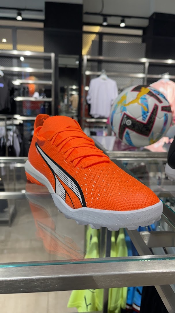 A close-up of an orange Puma soccer cleat with a soccer ball in the background.