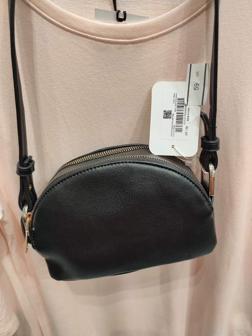 A black crossbody bag with a strap is displayed against a light pink garment.