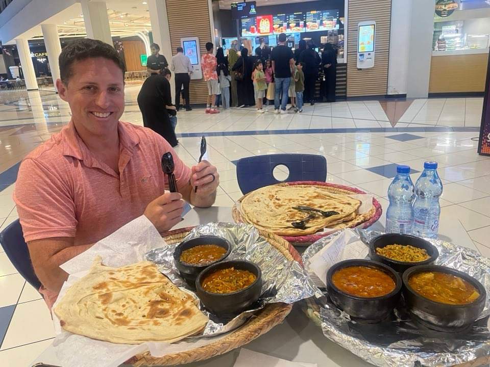 A man smiles at the camera with a table full of Indian food and naan bread.