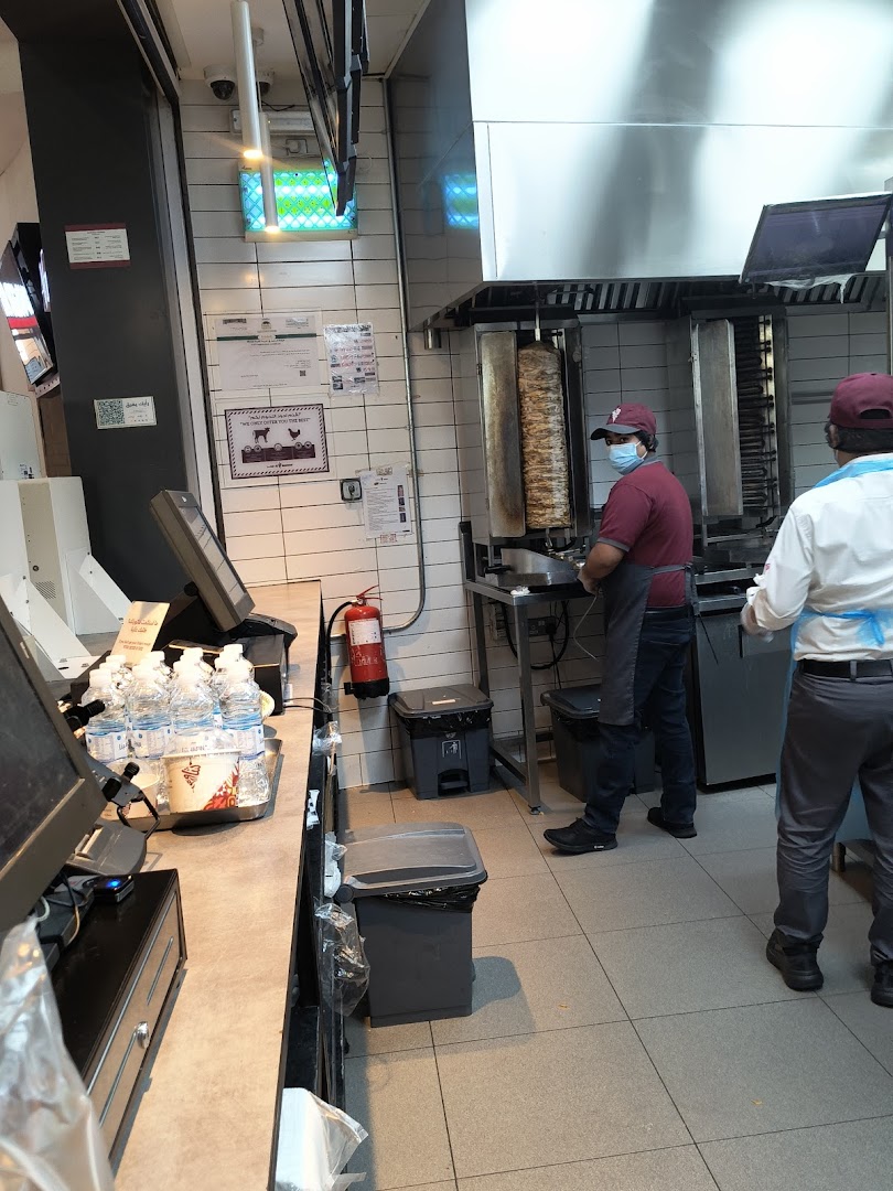 Two staff members preparing food behind a counter with shawarma grills and a cash register.
